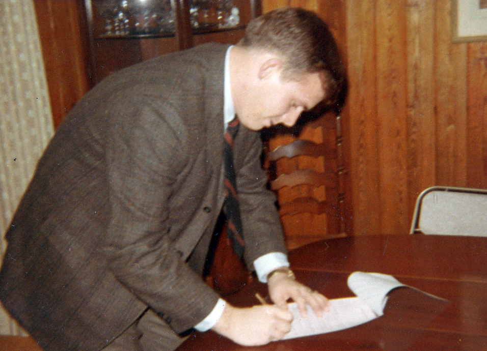 A young man wearing a brown suit signs a piece of paper on a dining table.