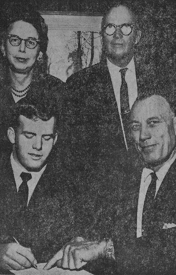 A newspaper photo shows a young man signing a contract while two men and a woman watch.