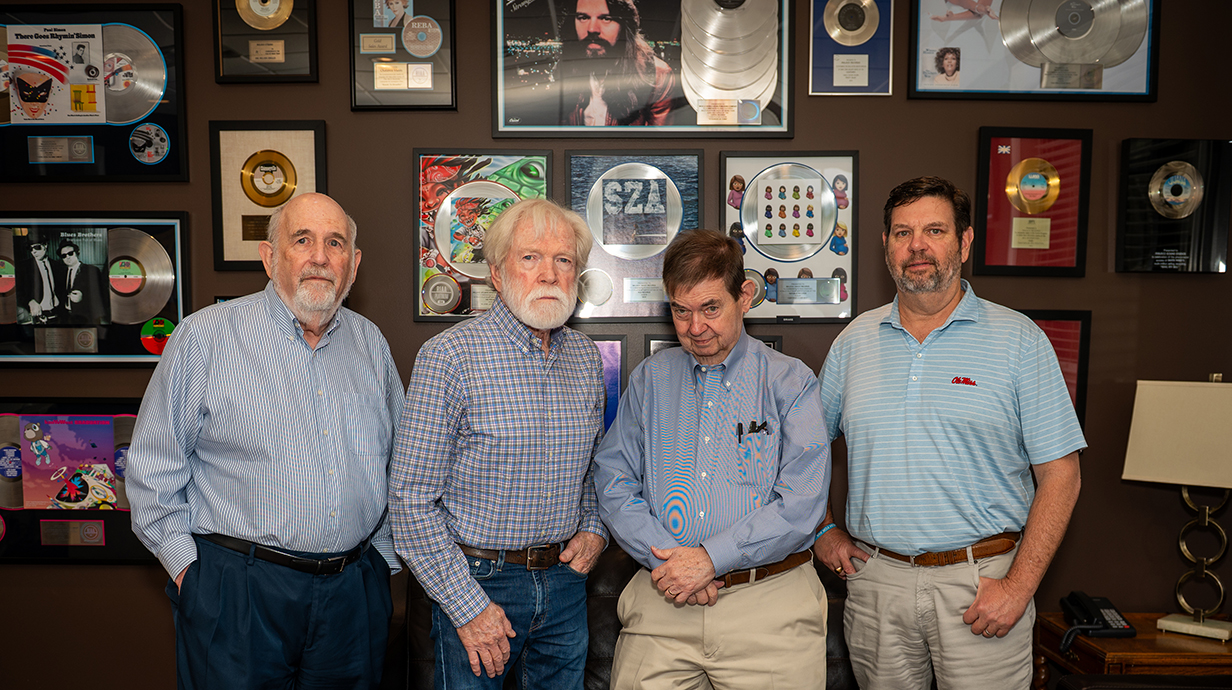 Four men stand in front of a wall lined with framed record albums.