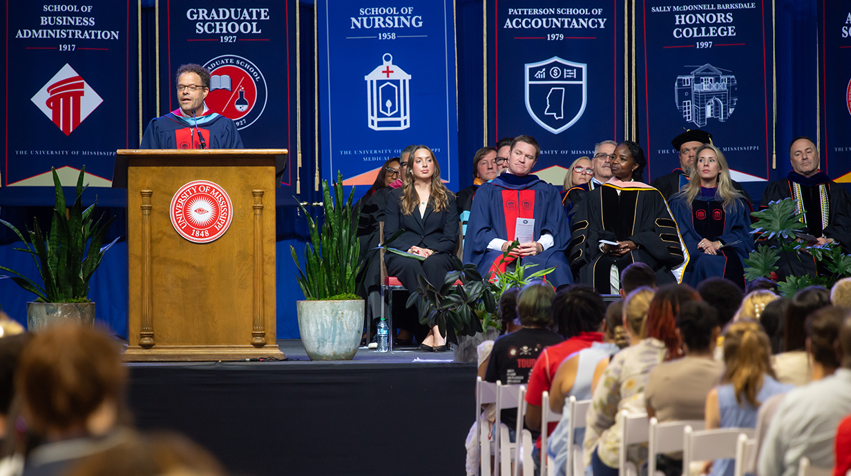 A man standing at a podium on a stage speaks to a crowd of young people seated in chairs on an arena floor.