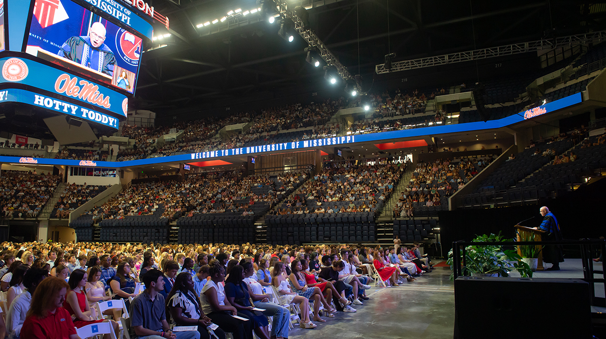 A man on a stage speaks to a large crowd of people seated in an arena.