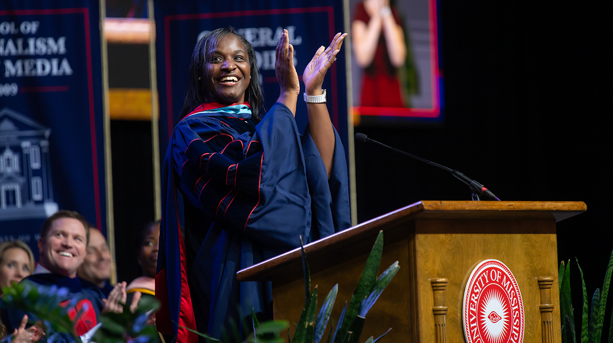 A woman wearing academic regalia at a podium claps while looking out on a crowd in an arena.