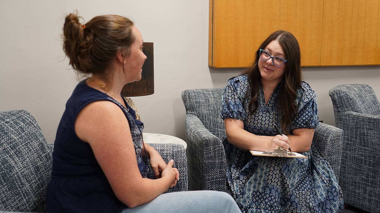 A woman holding a clipboard talks with a younger woman while they sit in a cozy reception area.