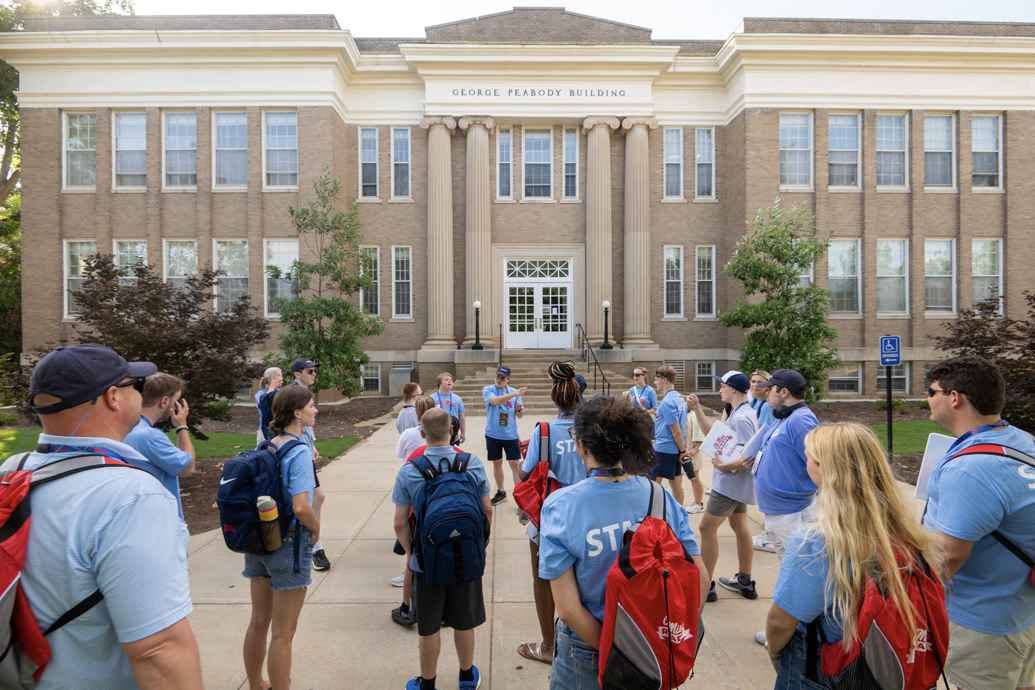 Man in polo shirt talks to a group of students outdoors near steps.
