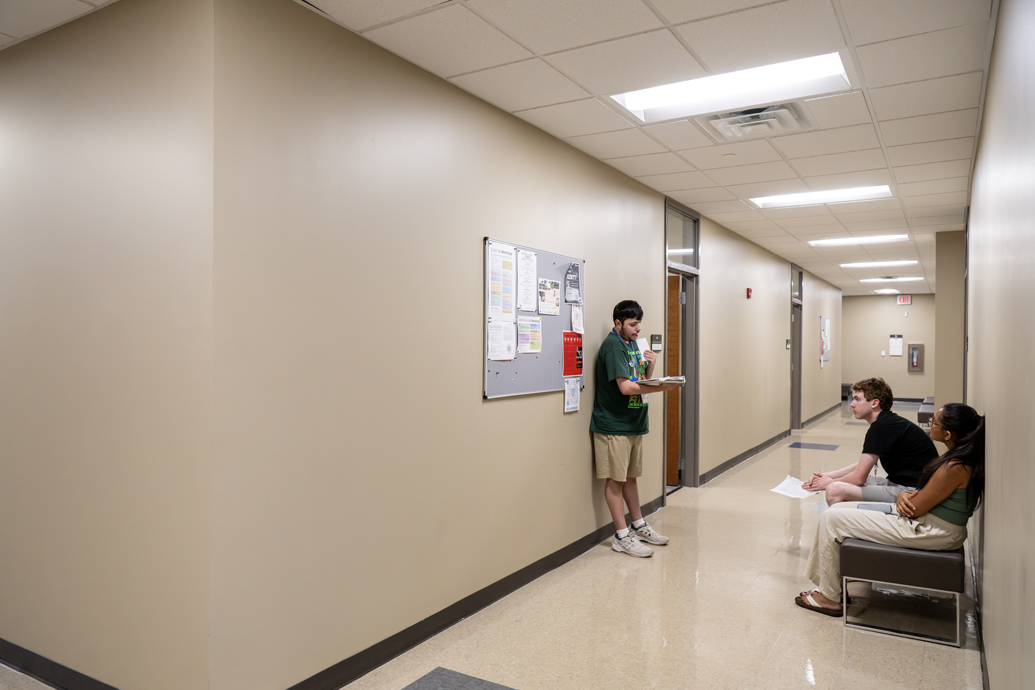 Two young men practice speeches with a woman in the hallway outside a classroom.