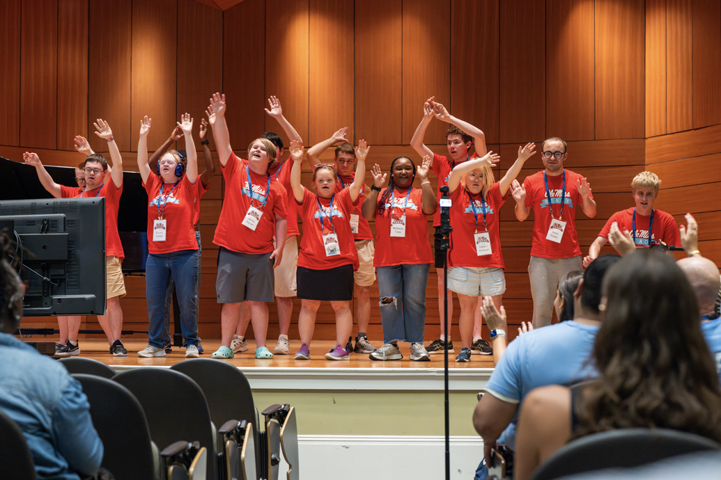 Group of students in red shirts perform onstage with hands raised singing.