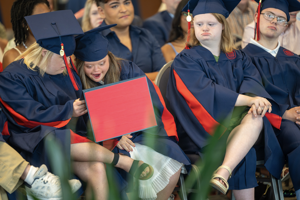 Young woman reacts with a hand on her chest as two others look on, holding certificates.