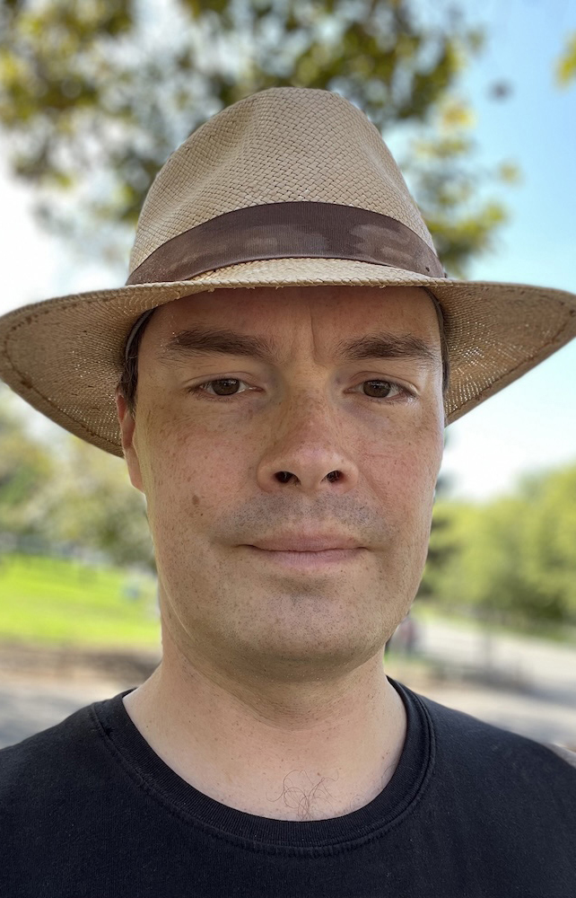 Headshot of a man wearing a black T-shirt and a fedora.