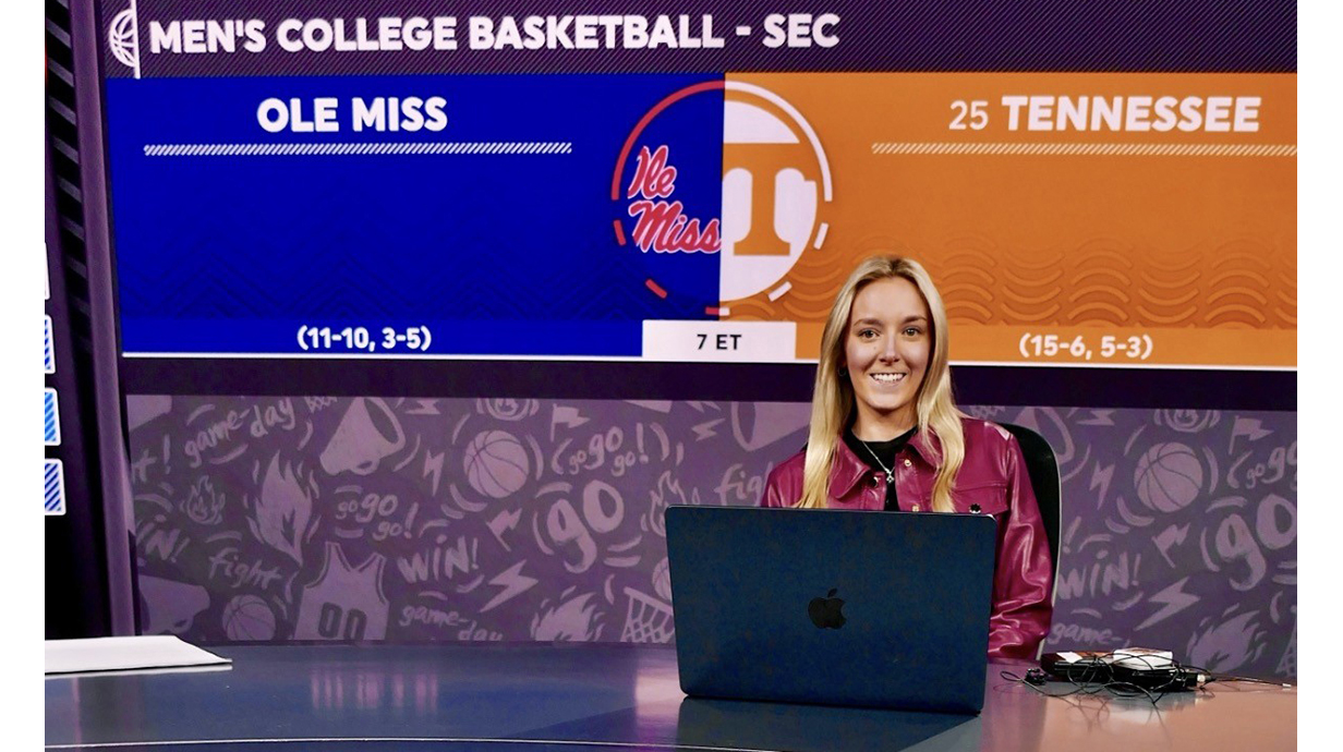 A young woman sits at a sports broadcast desk with Ole Miss and Tennessee logos projected behind her.
