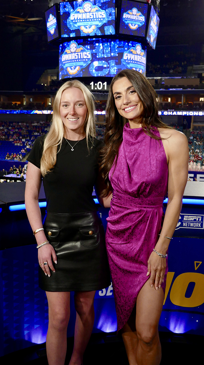 Two women stand in front of a broadcast desk in an arena with signs for a gymnastics competition.