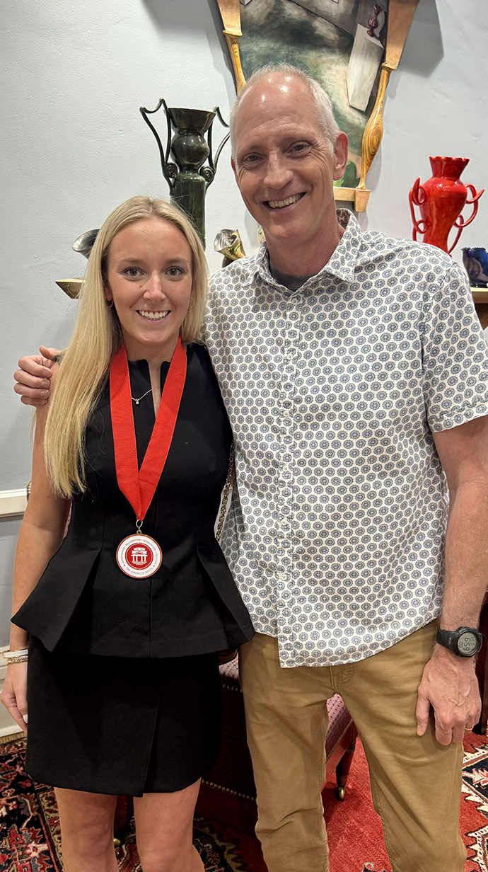 A woman wearing an award medal around her neck and a man stand in front of a colorful sculpture.