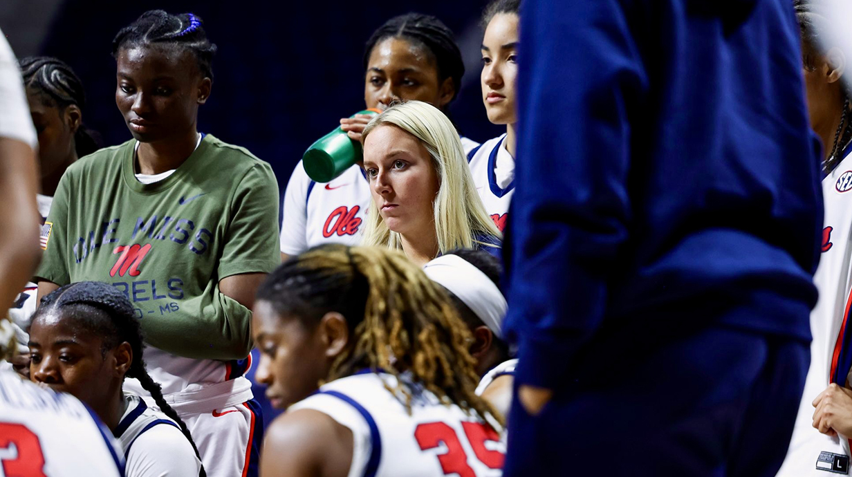 Several young women listen intently to a speaker not in the frame at a basketball arena.
