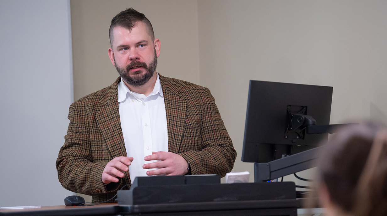 A man wearing a brown plaid jacket over a white dress shirt talks and gestures at the front of a classroom.