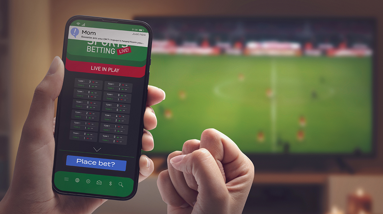 A man's hands hold a cellphone displaying a sports betting site  in front of a television tuned to a soccer game.