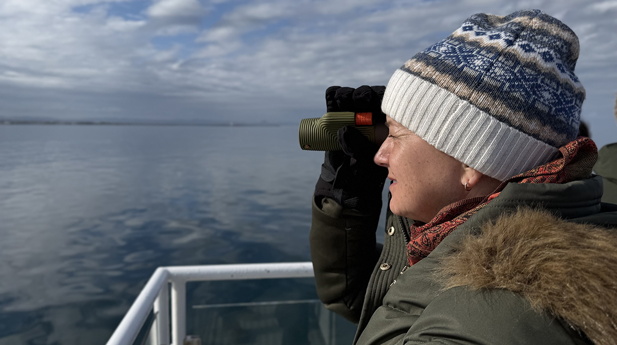 A woman wearing a ski hat and heavy coat looks through a monocular across a stretch of ocean.