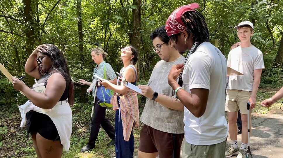 A group of young people walk through a wooded area.