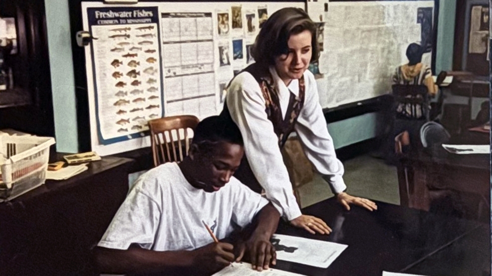 A teacher, standing, points out something to a seated student in a classroom.