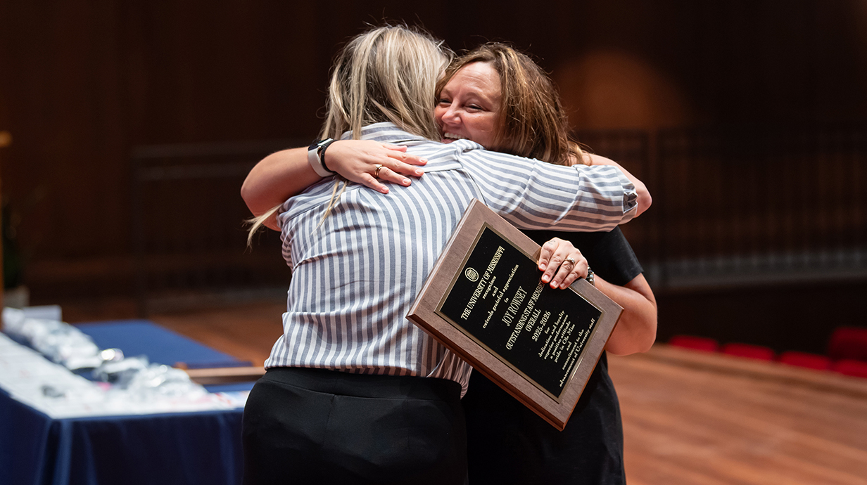 A woman holding an award plaque hugs another woman while standing on a stage in a theater.