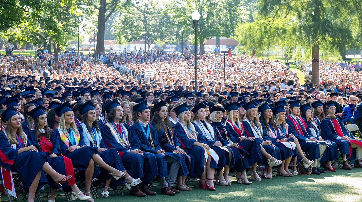 A large group of people wearing graduation regalia sit in chairs underneath towering trees in a park.
