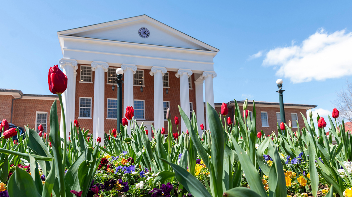 Colorful flowers bloom in front of a large brick building with white columns on its facade.