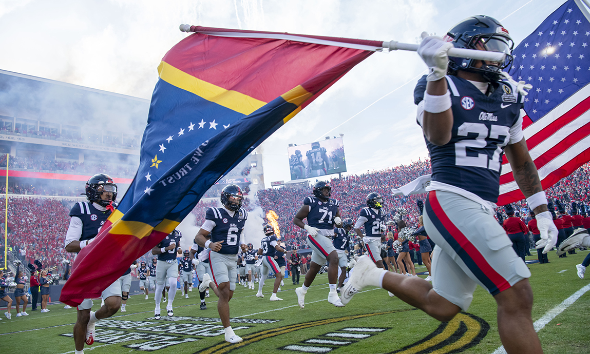 Football players and cheerleaders run onto a field in a stadium carrying flags.