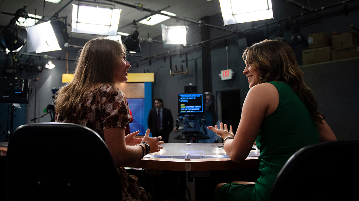 Two young women talk while sitting at a desk in a TV news studio while a young man stands by a teleprompter.