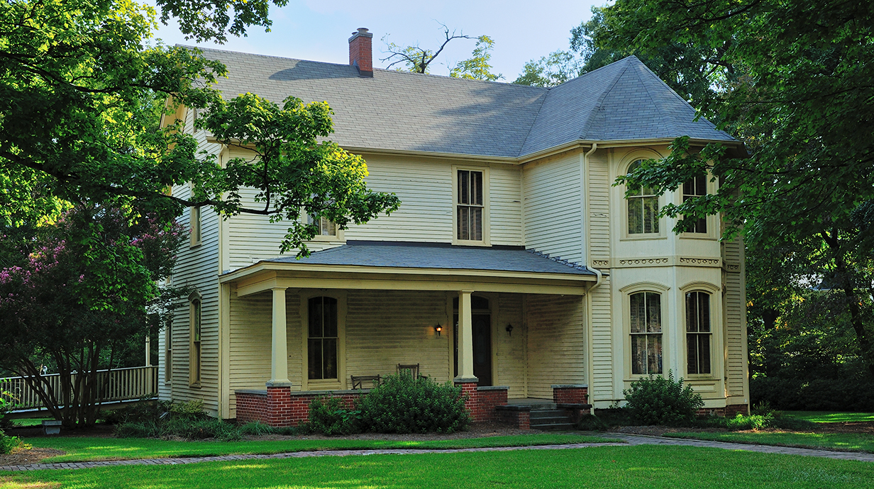 A yellow frame two-story house sits on a grassy lot surrounded by towering trees.