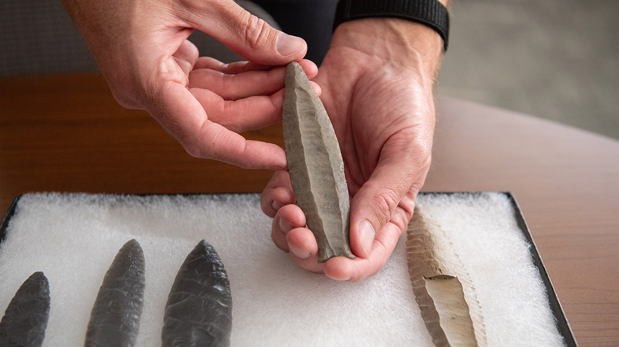 A man's hands hold a stone spear point beside a collection of other spear tips on a white board.