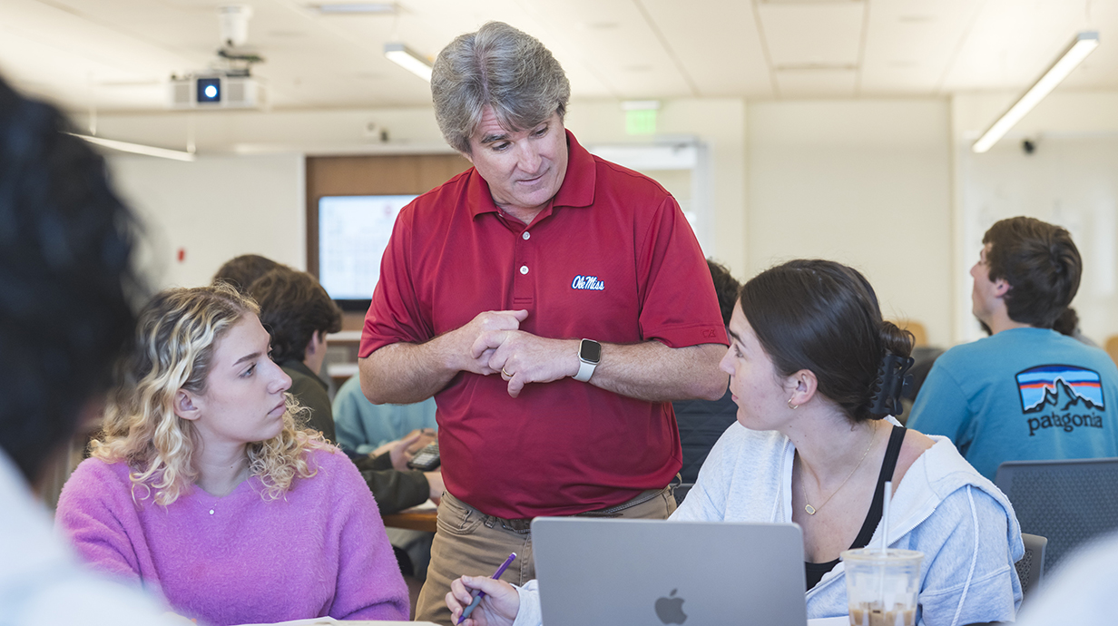A man wearing a red Ole Miss polo stands in a classroom and talks to two young women seated at a table.