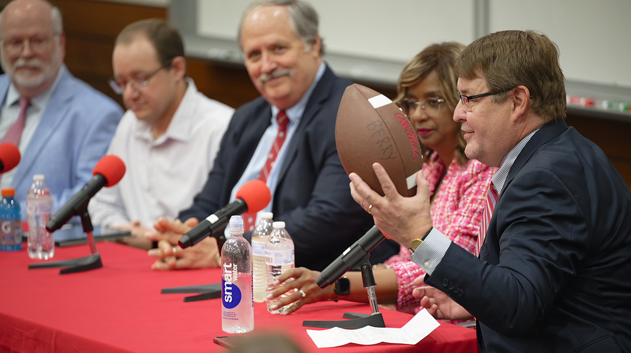 A man holding a football talks while sitting at one end of a table lined with three other men and a woman.