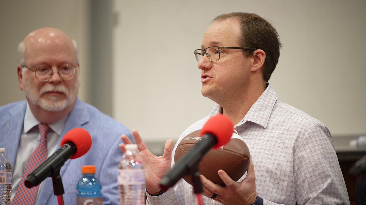 A man holding a football speaks into microphones while anotehr man watches.