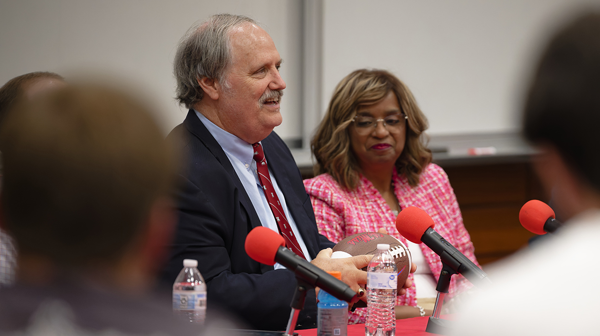 A man sitting at a table talks to people in a crowded auditorium as a woman watches.