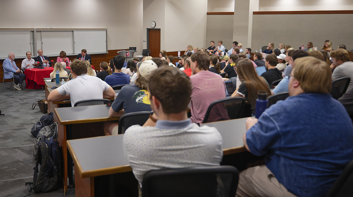 A crowd seated in an auditorium watches as five people seated at the front talk into microphones.