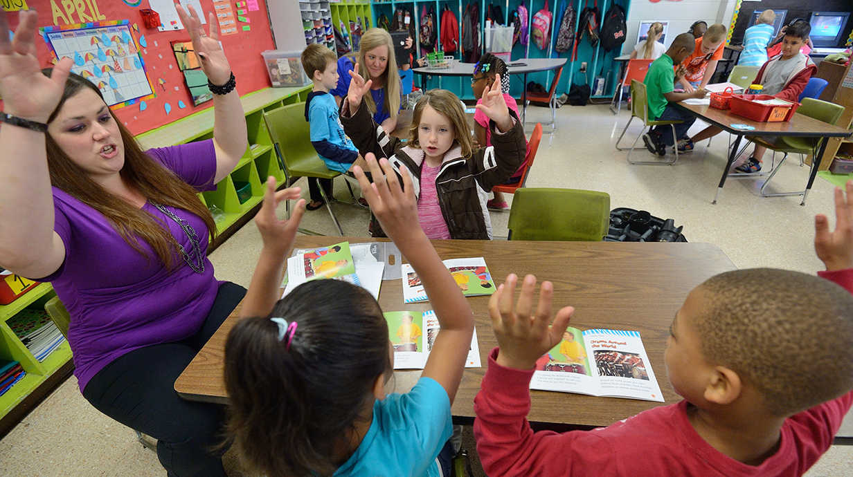 A young woman holds up her hands as she leads several small children seated around a low table in an activity in a classroom.