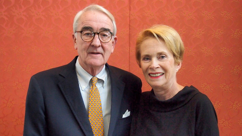 A man and a woman wearing dress clothes stand in front of a patterned red wall.