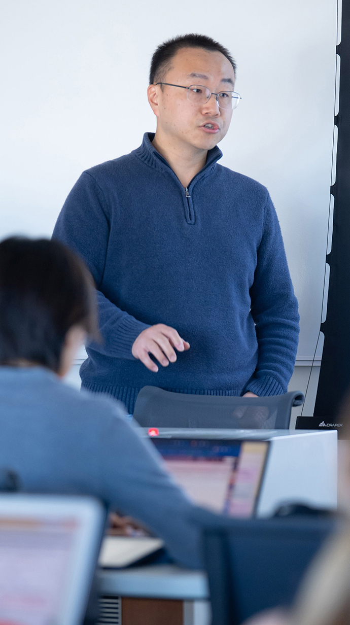A man wearing a blue sweater speaks from the front of a classroom.