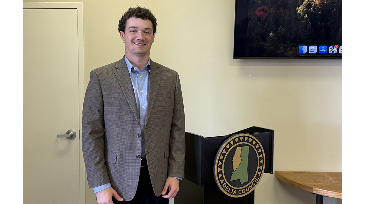 A young man wearing a brown suit stands by a lectern with a Delta Council plaque on the front.