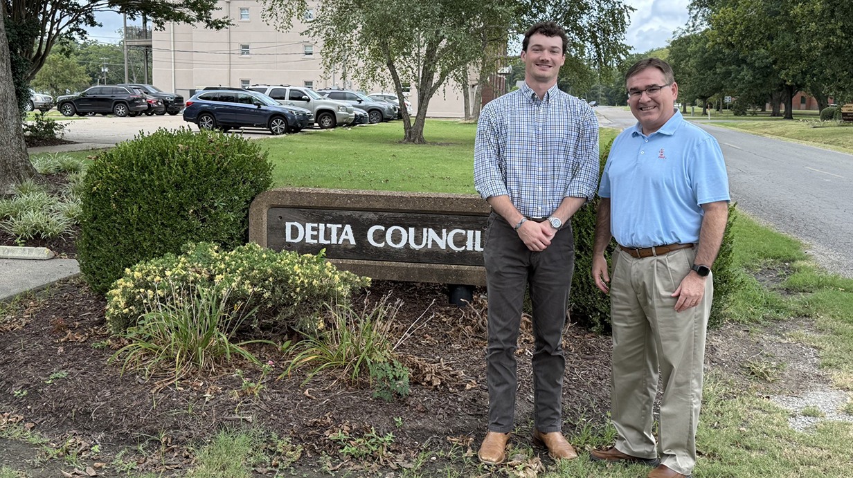 Two men stand in front of an landscaped area with a low sign reading 'Delta Council.'