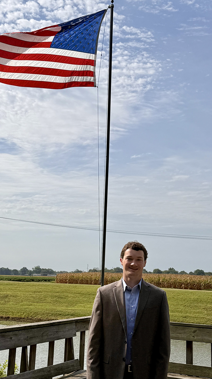 A young man stands in front of a flagpole flying an American flag.