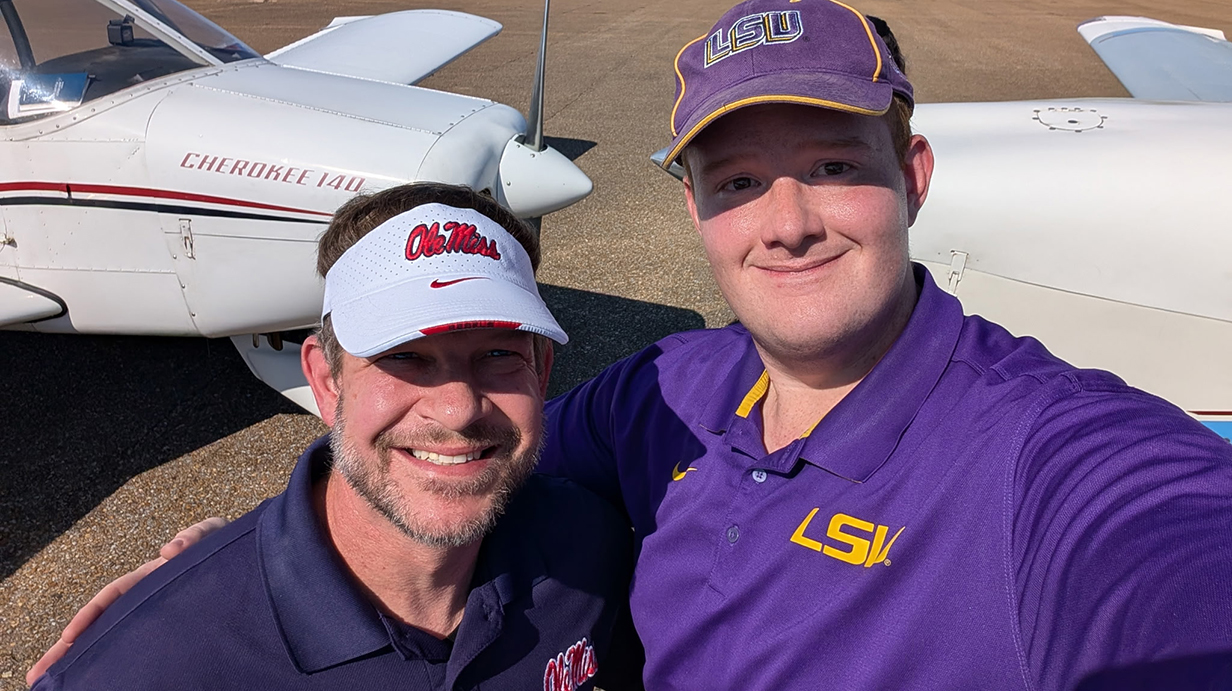 Two men, one wearing Ole Miss gear and the other dressed in LSU gear, take a selfie on an airport tarmac in front of two small airplanes.