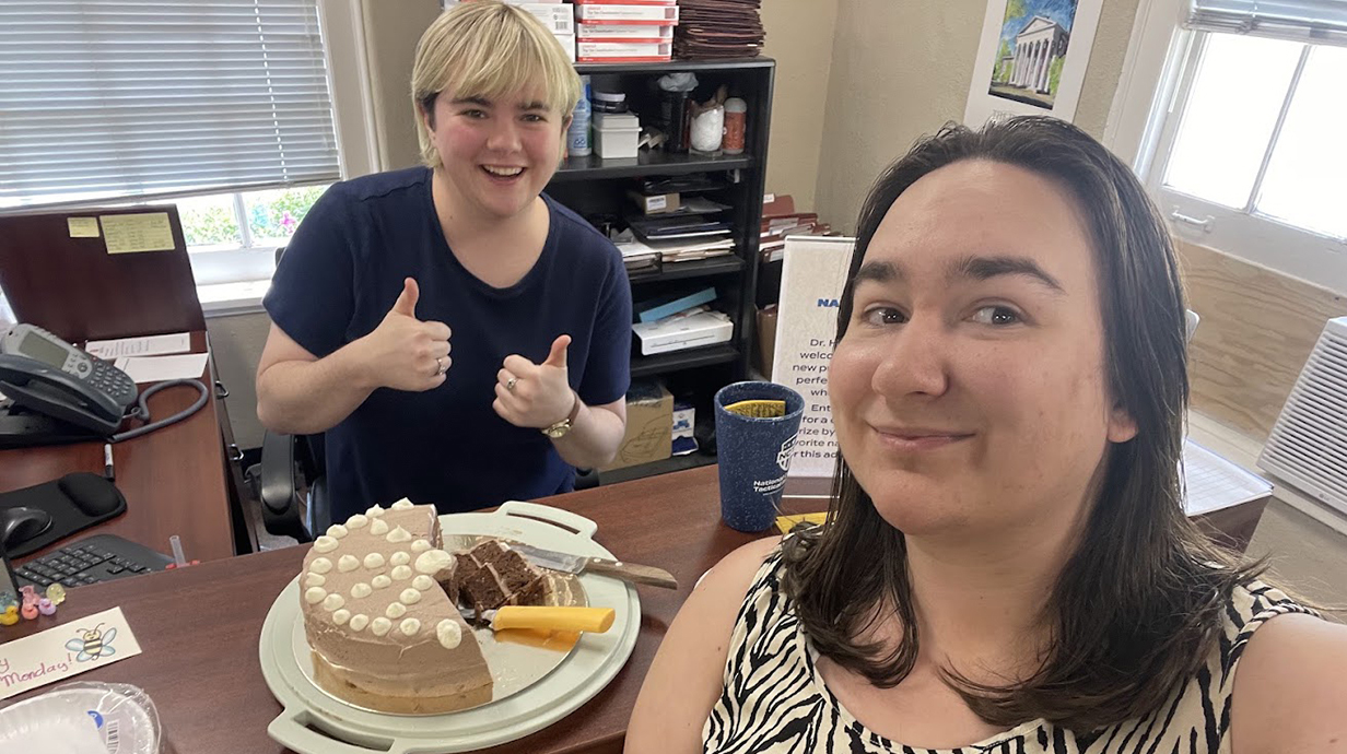 A woman gives a thumbs-up sign while standing behind a chocolate cake in an office as another woman smiles.