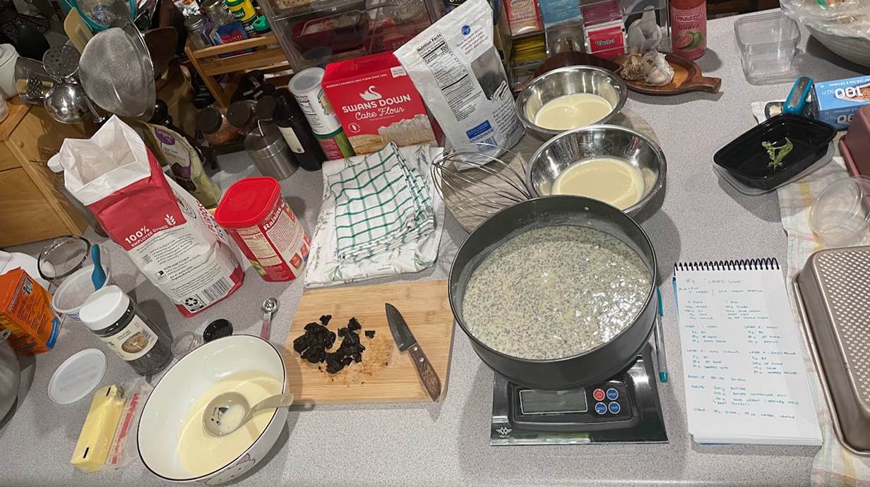 A kitchen counter is covered with ingredients, a food scale, a recipe and a pan of batter.
