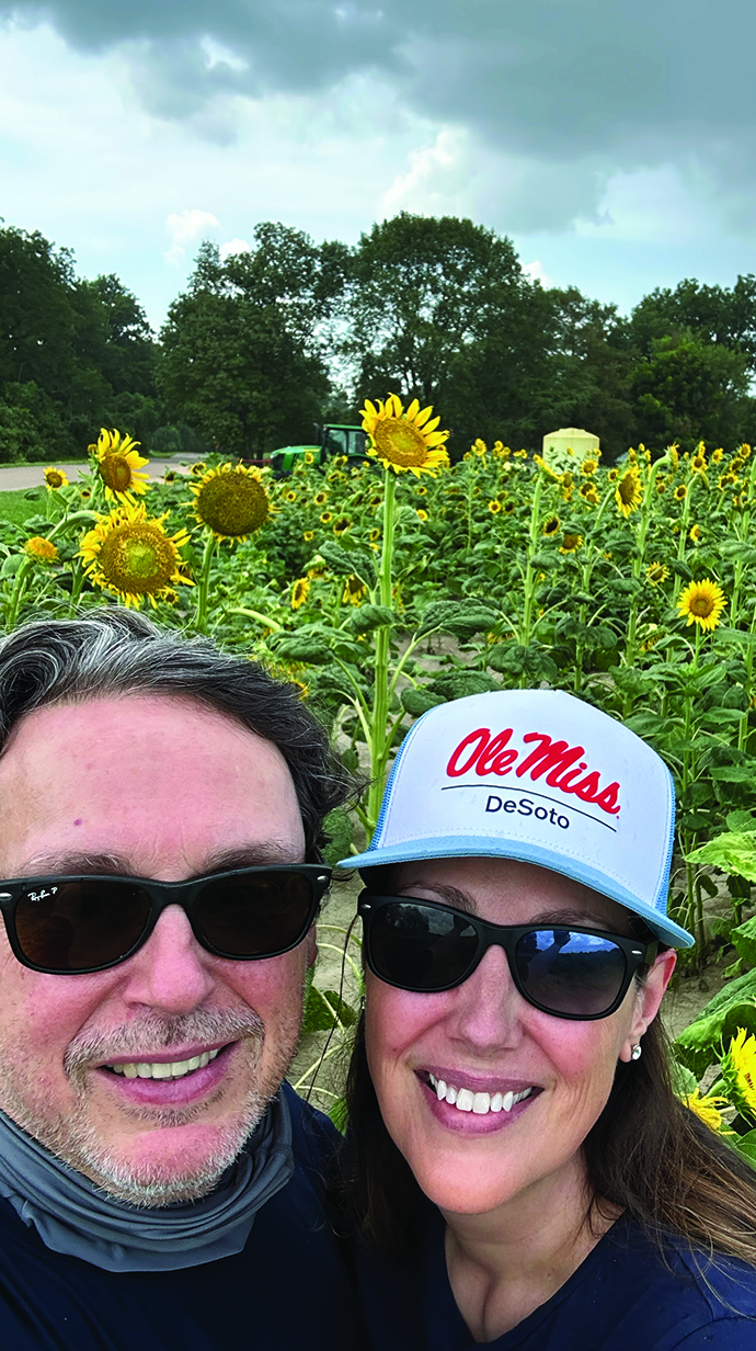 A man and a woman wearing an Ole Miss cap take a selfie in front of a field of colorful sunflowers.