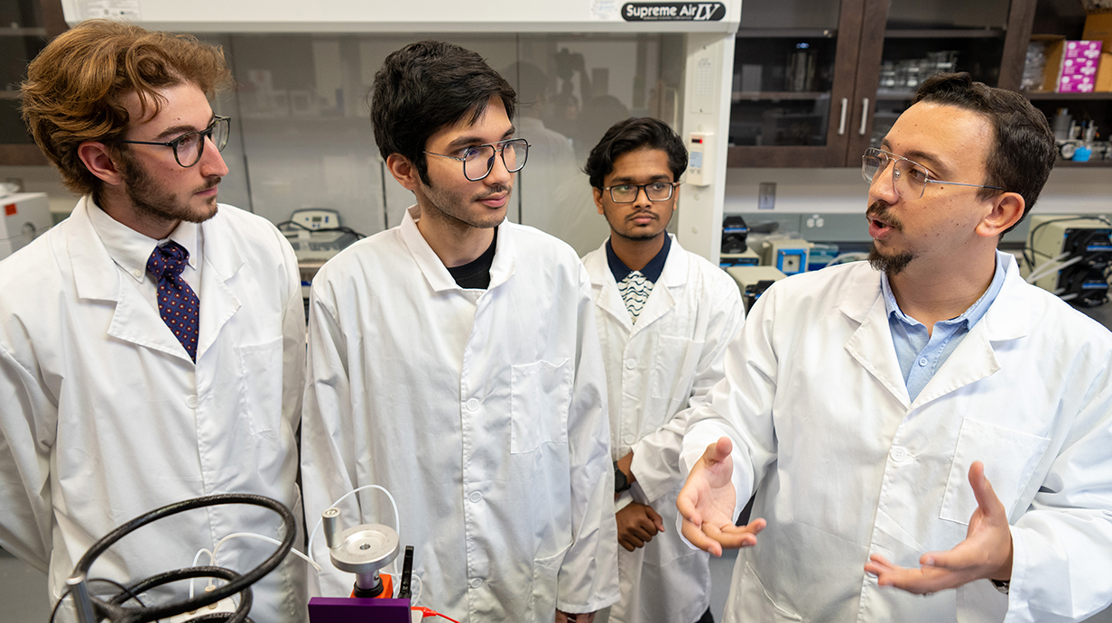 A man wearing a white lab coat talks with three young men, all wearing lab coats, in a laboratory.