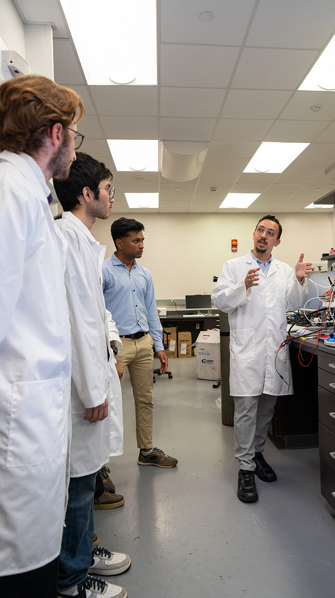 A man wearing a white lab coat talks with several young men wearing lab coats in a laboratory.