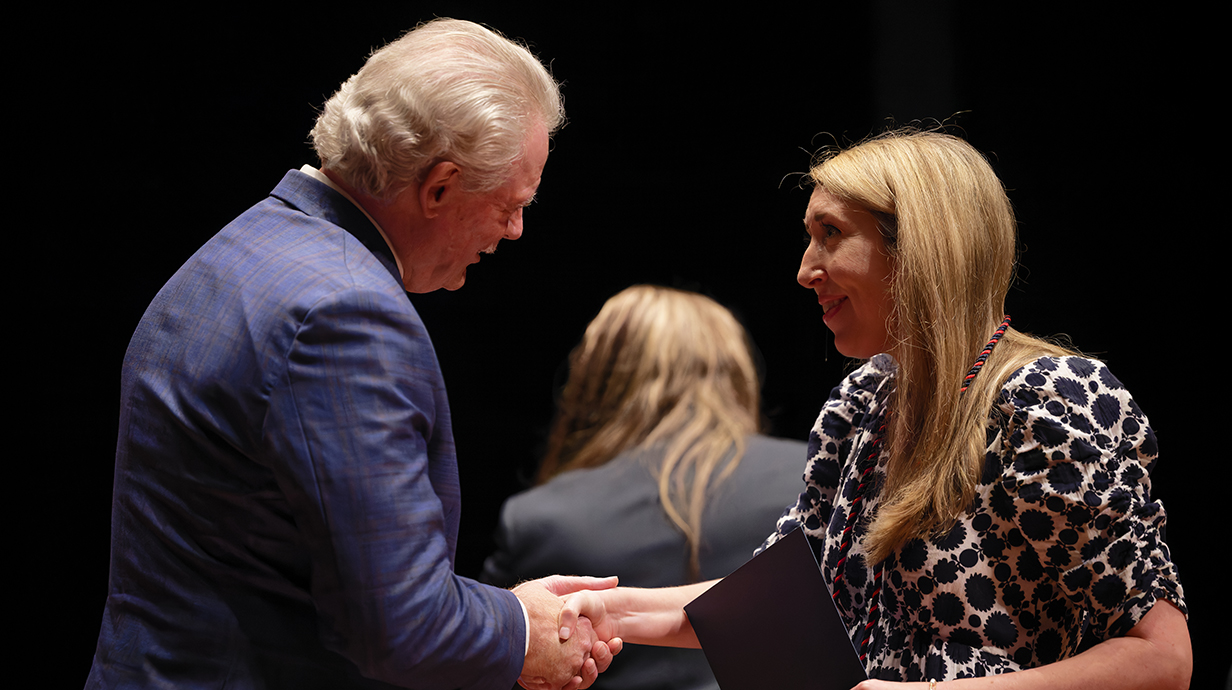 A man wearing a blue suit shakes a woman's hand and presents her with a certificate on a stage.