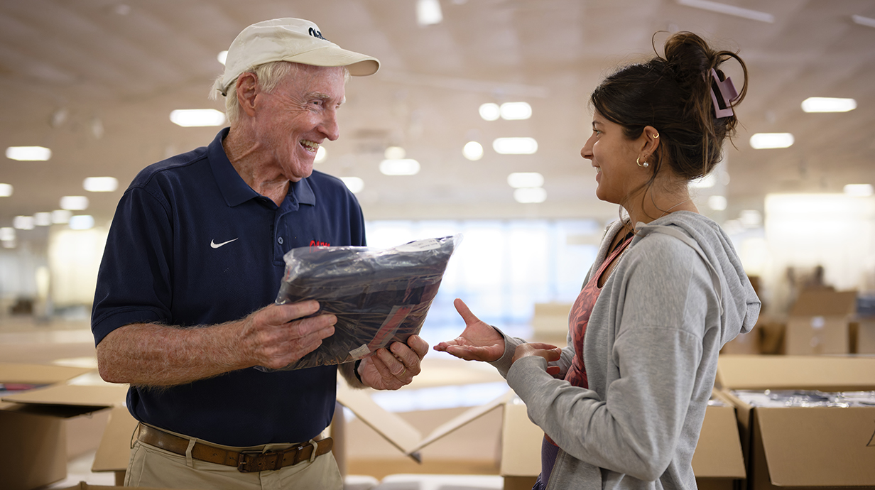 A man wearing a blue polo shirt and a baseball cap hands a package of clothing to a young woman.