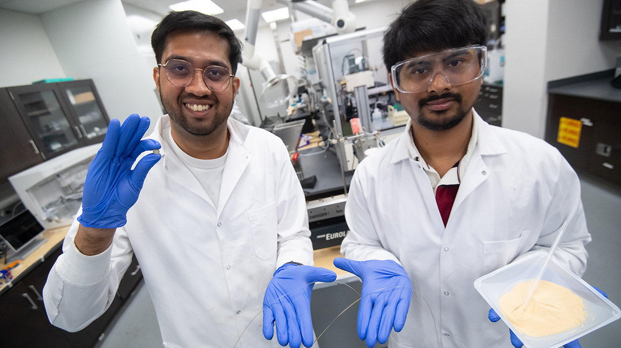 two young men wearing white lab coats, safety glasses and blue gloves hold a translucent filament in a lab.