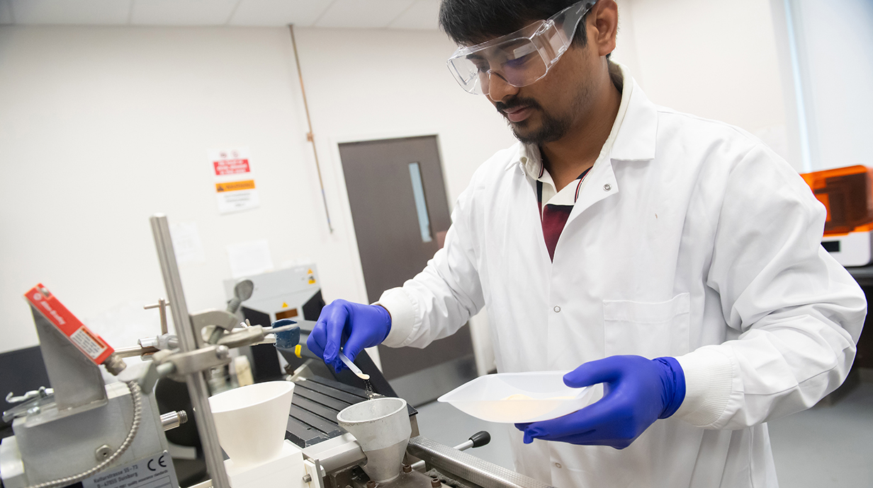 A young man wearing a white lab coat and blue gloves pour a small scoop of powder into a macine in a lab.
