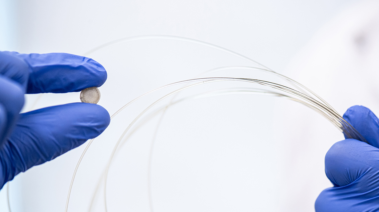 Gloved fingers hold a small tablet and a translucent filament.